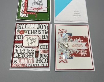 Puede incluir: Cuatro tarjetas de Navidad hechas a mano con diferentes diseños. La primera tarjeta tiene un fondo verde con papel estampado en rojo y blanco y el texto "Joyful Greetings". La segunda tarjeta tiene un fondo azul con un triángulo blanco y el texto "As many snowflakes on a winter day can told". La tercera tarjeta tiene un fondo estampado en rojo y blanco con el texto "Joy Love Peace" y otras palabras navideñas. La cuarta tarjeta tiene un fondo estampado en rojo y blanco con un copo de nieve plateado y el texto "Happiest of Holidays".
