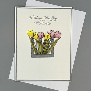 Puede incluir: Una tarjeta blanca con un borde gris y el texto "Wishing You Joy At Easter" en escritura cursiva negra. La tarjeta presenta un marco gris con un borde cosido y un ramo de tulipanes amarillos y rosas.
