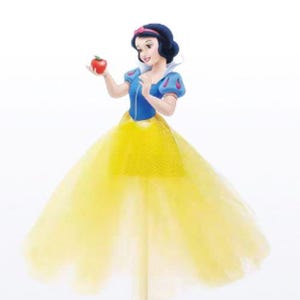 May include: A Snow White figurine holding a red apple. The figure wears a blue top with puffy sleeves, a yellow skirt, and a red headband. The figurine is a cake topper.