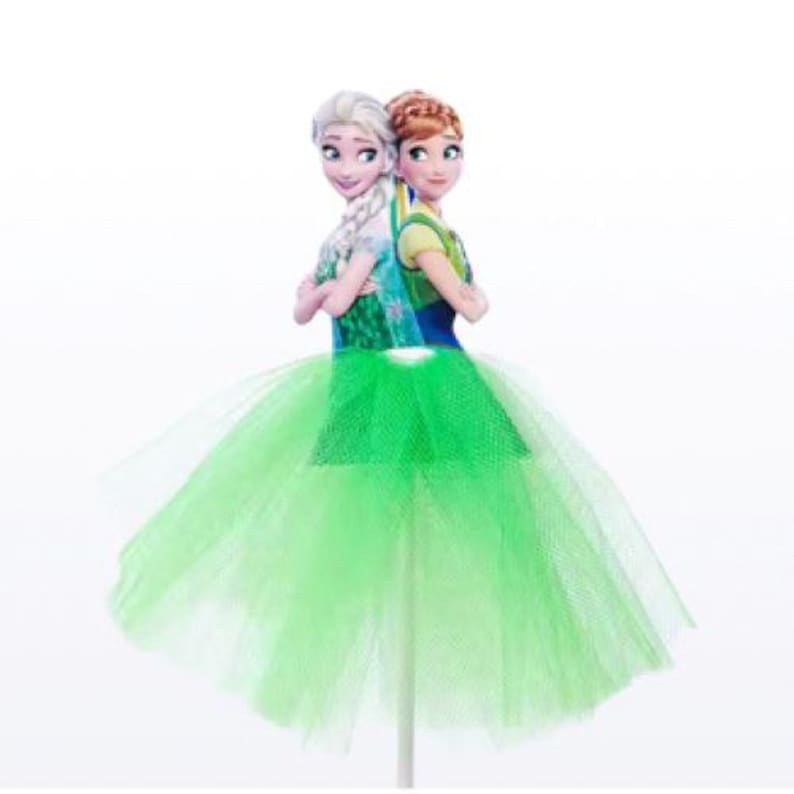 May include: A cake topper featuring two cartoon figures, one with blonde hair and the other with auburn hair, standing back-to-back. They are wearing dresses and are set atop a bright green tulle skirt. The cake topper is on a white stick.