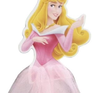 May include: A die-cut image of Princess Aurora from Disney's Sleeping Beauty. She is depicted with long, flowing blonde hair, wearing a pink gown with a tulle skirt and a gold crown. The image has a white border.