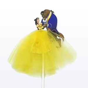 May include: A yellow tulle skirt with a cartoon cutout of Belle and the Beast dancing. The figures are on a stick, likely a cake topper. The skirt is a bright, sunny yellow.