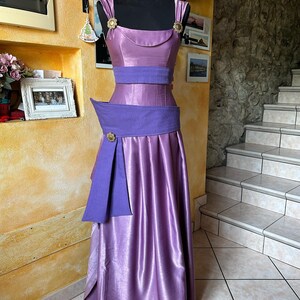 May include: A lilac formal dress displayed on a black mannequin. The dress has a fitted bodice with a scoop neckline, a wide purple belt, and a flowing skirt. Gold floral accents are on the straps and belt. The dress is made of a shiny, satin-like fabric.