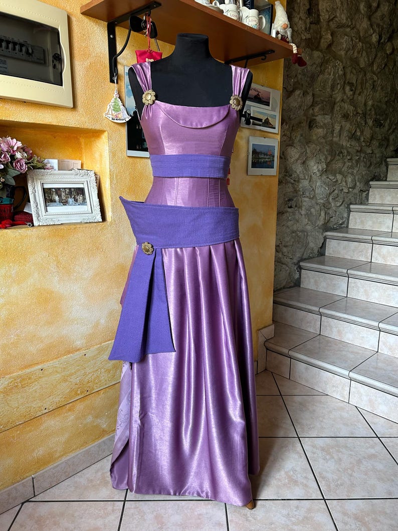 May include: A lilac formal dress displayed on a black mannequin. The dress has a fitted bodice with a scoop neckline, a wide purple belt, and a flowing skirt. Gold floral accents are on the straps and belt. The dress is made of a shiny, satin-like fabric.