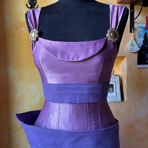 May include: A lavender satin dress on a black mannequin. The dress has a square neckline, wide straps, and a cinched waist with a textured belt. Bronze-coloured clasps decorate the straps and skirt. The skirt is pleated. The dress is a vintage style.
