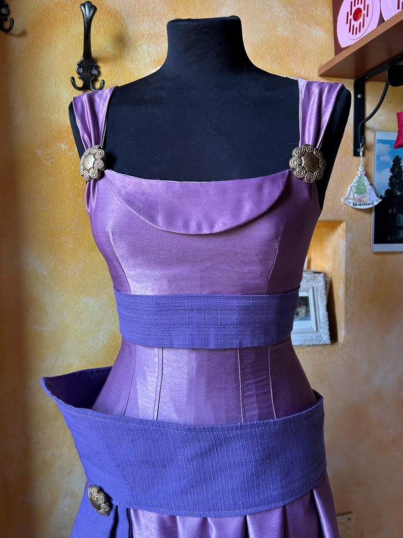 May include: A lavender satin dress on a black mannequin. The dress has a square neckline, wide straps, and a cinched waist with a textured belt. Bronze-coloured clasps decorate the straps and skirt. The skirt is pleated. The dress is a vintage style.