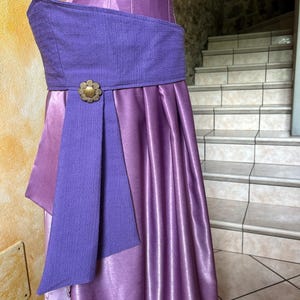May include: A two-piece purple dress with a textured bodice and a flowing skirt. The dress features a wide, textured purple belt with a gold-coloured floral buckle. The skirt has vertical pleats, and the overall design is elegant and formal.