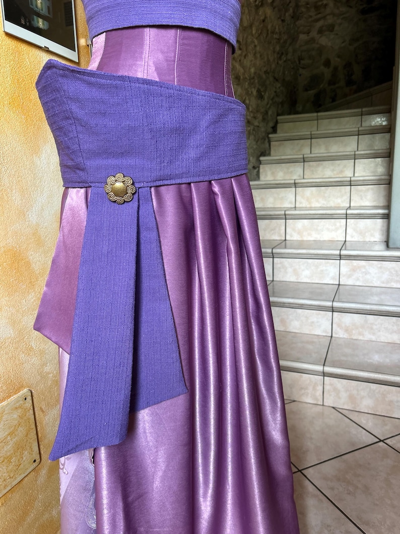 May include: A two-piece purple dress with a textured bodice and a flowing skirt. The dress features a wide, textured purple belt with a gold-coloured floral buckle. The skirt has vertical pleats, and the overall design is elegant and formal.