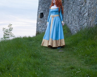 May include: A woman in a blue and gold dress stands on a grassy hill near a stone castle.