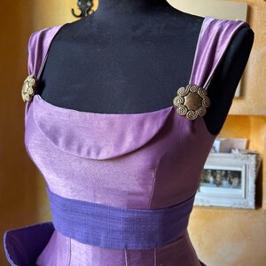 May include: A lilac dress with a scoop neckline and a wide, dark purple belt. The dress has decorative gold-toned clasps on the shoulder straps. The dress is displayed on a black mannequin.