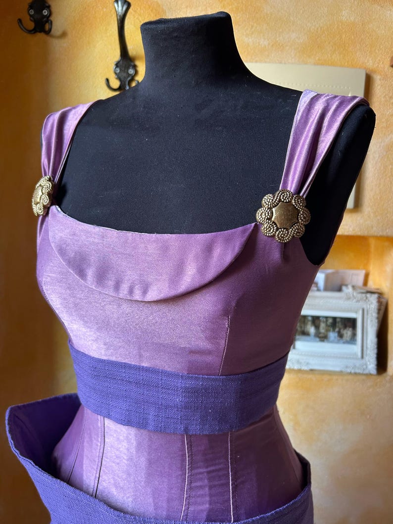May include: A lilac dress with a scoop neckline and a wide, dark purple belt. The dress has decorative gold-toned clasps on the shoulder straps. The dress is displayed on a black mannequin.