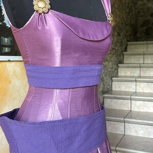 May include: A lilac dress with a fitted bodice and a pleated skirt. It has a wide, textured purple belt and gold-toned embellishments. The dress is displayed on a black mannequin.