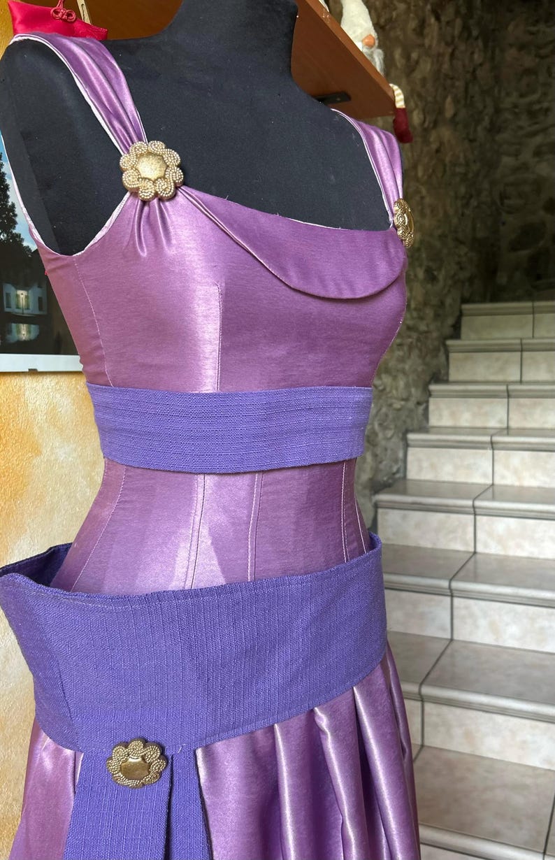 May include: A lilac dress with a fitted bodice and a pleated skirt. It has a wide, textured purple belt and gold-toned embellishments. The dress is displayed on a black mannequin.