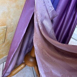 May include: Draped fabric in shades of purple and lavender. The fabric has a silky sheen and a subtle paisley pattern. The folds of the fabric are visible, creating depth and texture. The fabric is draped over a wooden stand.