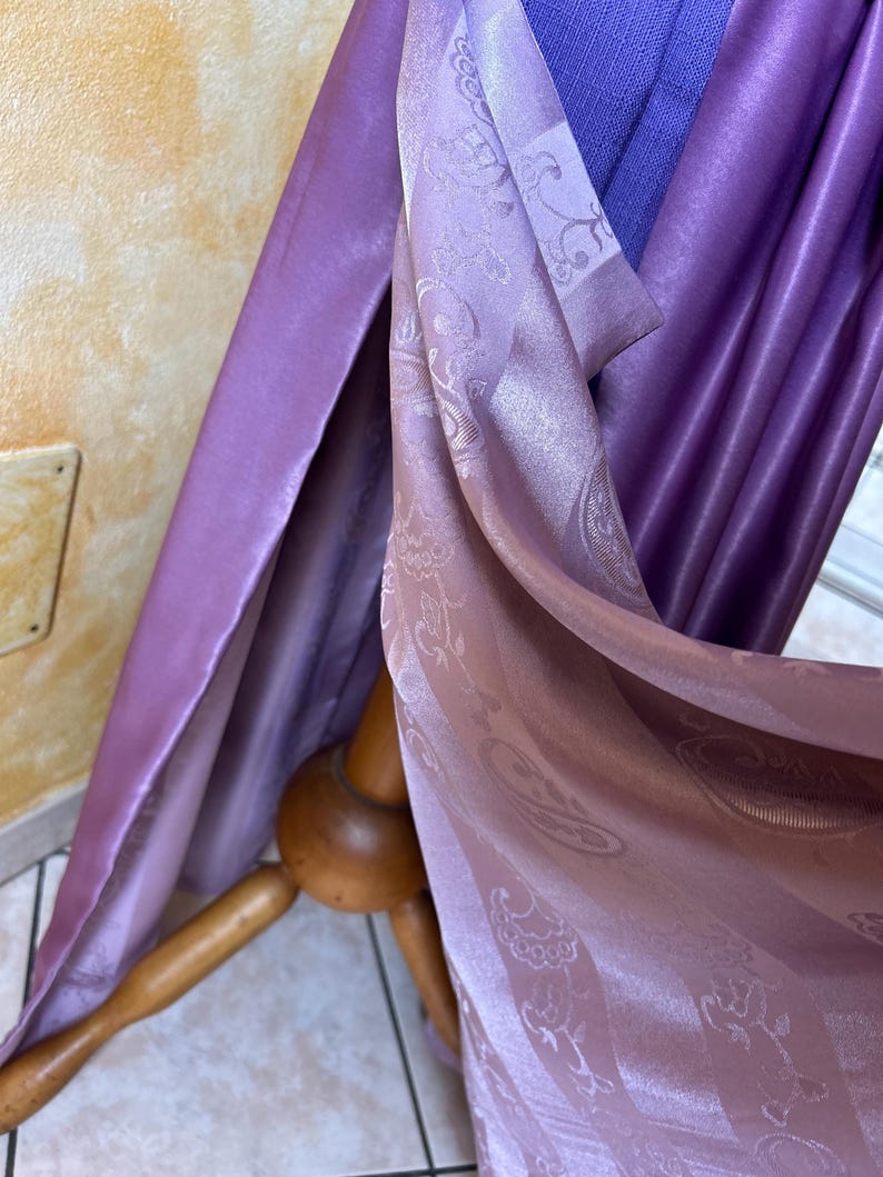 May include: Draped fabric in shades of purple and lavender. The fabric has a silky sheen and a subtle paisley pattern. The folds of the fabric are visible, creating depth and texture. The fabric is draped over a wooden stand.