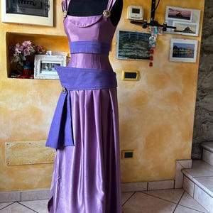 May include: A lilac formal gown on a black mannequin. The dress has a fitted bodice and a wide, pleated skirt. The bodice has a scoop neckline and is accented with a wide, purple belt and gold-coloured embellishments.