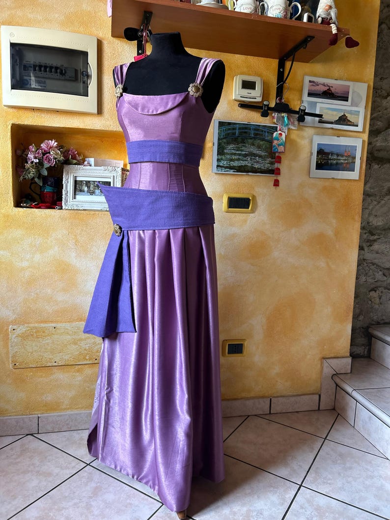 May include: A lilac formal gown on a black mannequin. The dress has a fitted bodice and a wide, pleated skirt. The bodice has a scoop neckline and is accented with a wide, purple belt and gold-coloured embellishments.