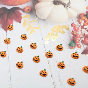 May include: A collection of 15 small orange pumpkin-shaped Halloween decorations with black eyes and a toothy grin. The pumpkins are arranged on a white wooden surface.