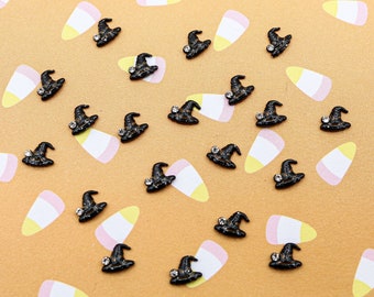 May include: A collection of 20 black witch hat charms with a single rhinestone on each hat. The charms are scattered on a yellow and white candy corn patterned background.