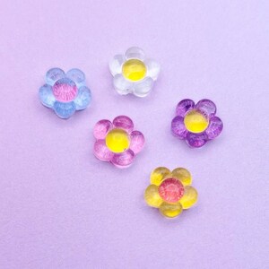 May include: Five colorful flower-shaped embellishments. Each flower has a translucent petal design with a yellow center. The colors include blue, white, pink, purple, and yellow. The background is a solid lavender color.