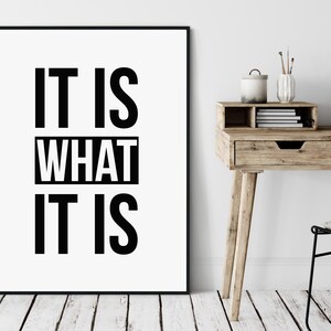 May include: A framed print with the text "IT IS WHAT IT IS" in bold black letters on a white background. The print is displayed on a white wall above a wooden desk with a laptop, books, and decorative items.