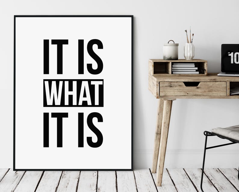 May include: A framed print with the text "IT IS WHAT IT IS" in bold black letters on a white background. The print is displayed on a white wall above a wooden desk with a laptop, books, and decorative items.