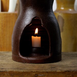 May include: A brown ceramic candle holder with a cutout design and a lit candle inside. The holder has a handle and is sitting on a wooden surface.