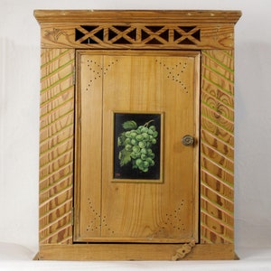 May include: A wooden cabinet with a single door and a painted image of a bunch of green grapes on the front. The cabinet has a decorative top with a lattice design and green and pink painted accents.