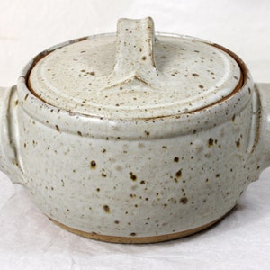 May include: A white ceramic casserole dish with a lid and two handles. The dish has a speckled glaze and a brown rim.
