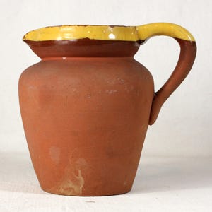 May include: A terracotta pitcher with a yellow rim and handle. The pitcher has a rounded body and a slightly flared lip.