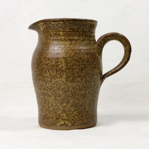 May include: A handmade, speckled brown ceramic jug with a curved handle and a pouring spout. The jug has a rustic, textured appearance, and is suitable for serving beverages. The jug is approximately 20 cm tall.