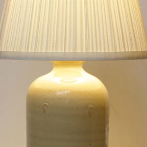 May include: A white ceramic lamp base with a pleated white lampshade. The lamp base has a textured surface and is shaped like a bottle.