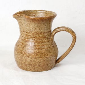 May include: A brown ceramic jug with a speckled glaze and a rounded body. The jug has a wide, slightly flared opening and a handle that curves up and over the top.