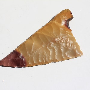 May include: A light brown and reddish-brown arrowhead made of flint. The arrowhead is triangular in shape with a pointed tip and a slightly curved base.
