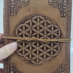 Op de afbeelding: Een bruin leren notitieboek met een geometrisch bloem des levens ontwerp in reliëf op de kaft. Het notitieboek heeft decoratieve hoekaccenten en is vastgemaakt met een bruin koord. Het leer heeft een verweerde uitstraling.