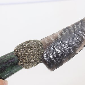 Puede incluir: Un cuchillo de pedernal gris oscuro con un mango verde y negro. El mango está envuelto en un material que se asemeja a una piedra. El cuchillo mide aproximadamente 15 cm de largo.