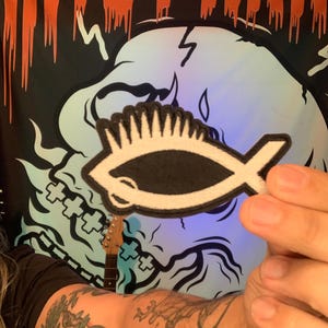 May include: A black and white fish-shaped patch with a distinctive design. The patch is held up in front of a black banner with orange text that reads "NATE PARA". A large, stylized skull graphic is visible in the background.