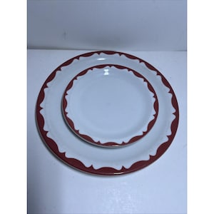 Puede incluir: Dos platos de cerámica blancos, uno anidado dentro del otro. Ambos platos tienen un diseño de borde festoneado en color rojo oscuro. La parte interior de los platos es blanca, creando un borde contrastante.