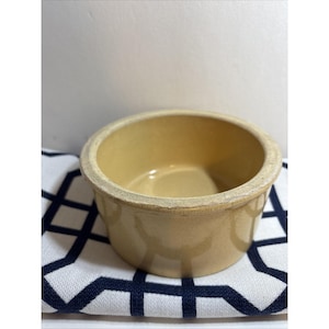 Puede incluir: Un cuenco pequeño y redondo de cerámica beige con un borde ligeramente rugoso. El cuenco está sobre un paño con estampado geométrico blanco y azul marino. El cuenco tiene un diseño sencillo y clásico.