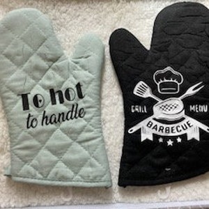 May include: A pair of oven gloves. One glove is green with the text "To hot to handle" in black. The other glove is black with a white design of a chef's hat, crossed utensils, and the text "Grill Barbecue Menu".