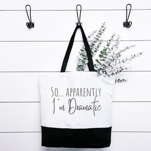 Könnte beinhalten: Eine weiße Canvas-Tragetasche mit schwarzen Henkeln und einem schwarzen Boden. Die Tasche hat den Text "SO...APPARENTLY I'm Dramatic" in Schwarz auf der Vorderseite aufgedruckt.