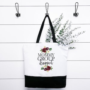 Könnte beinhalten: Weiße Canvas-Tragetasche mit schwarzen Henkeln und schwarzem Boden. Die Tasche hat ein Blumenmuster und den Text "Mommy Group Dropout" auf der Vorderseite.