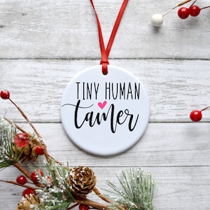 May include: White ceramic bauble with a red ribbon. The bauble has black text that reads "Tiny Human Tamer" with a pink heart.