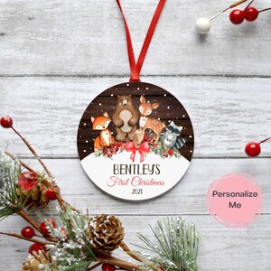 Puede incluir: Un adorno navide&ntilde;o redondo y blanco con un fondo de madera marr&oacute;n que presenta un zorro, un oso, un ciervo, un b&uacute;ho y un mapache. El adorno tiene el texto "Bentley's First Christmas 2021" impreso en &eacute;l.