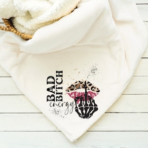 Può includere: Coperta bianca con una stampa grafica nera e dorata di labbra con stampa leopardata con una mano scheletrica che fa il dito medio. Il testo "BAD BITCH energy" è stampato in nero.
