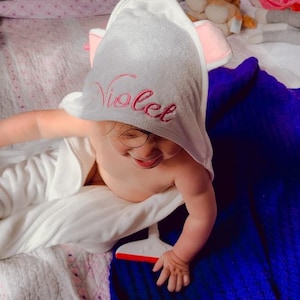 Op de afbeelding: Een witte badjas met een capuchon met roze oren en de naam "Violet" geborduurd op de voorkant. De badjas wordt gedragen door een kind dat op een blauwe en roze deken zit.