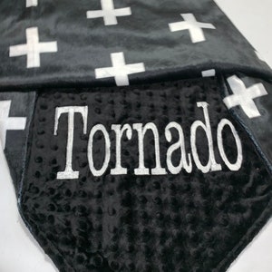 May include: A black and white baby blanket with a white cross pattern on one side and a black minky fabric with the name "Tornado" embroidered in white on the other side.