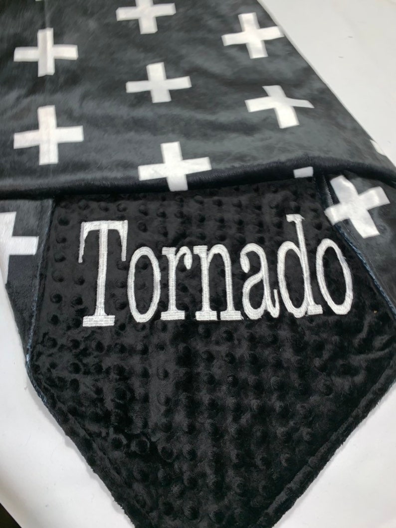 May include: A black and white baby blanket with a white cross pattern on one side and a black minky fabric with the name "Tornado" embroidered in white on the other side.