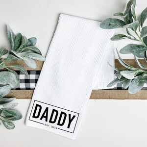 May include: White kitchen towel with black text that reads "Daddy Est. 2024".