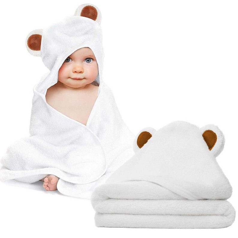 Op de afbeelding: Witte badcape met bruine berenoren voor een baby. De badcape is gevouwen en wordt getoond met een baby die de badcape draagt.